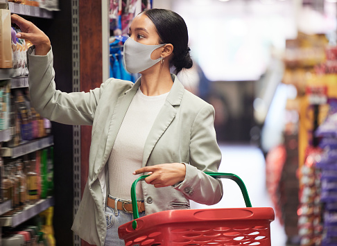 Supermarket, shopping and customer with covid face mask in retail store for food, groceries or product from shelf. Young woman with basket during inflation price increase or sales choice on grocery