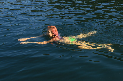 A Native woman doing the breast stroke while swimming in a lake in Autumn. She is wearing a red bikini top and green bottom.