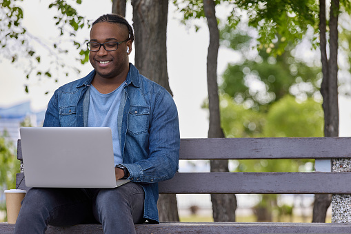 Black man traveler rests on a bench after a long route. Cute African guy smiling works online on the fresh thanks to modern technology. Digital technologies of online work
