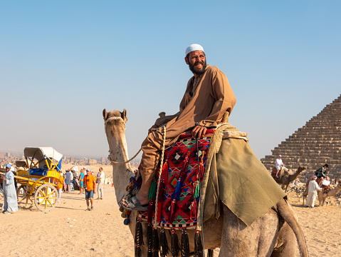 Giza, Cairo, Egypt - September 30, 2021: Egyptian in national dress sits astride a camel and smiles. Tourists walk and take pictures near the Egyptian pyramid.