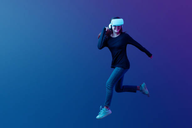 Young Asian woman wearing VR headset with experience playing video game and jumping levitating in the air on futuristic purple cyberpunk neon light background. Young Asian woman wearing VR headset with experience playing video game and jumping levitating in the air on futuristic purple cyberpunk neon light background. Metaverse technology concept. playing alone stock pictures, royalty-free photos & images