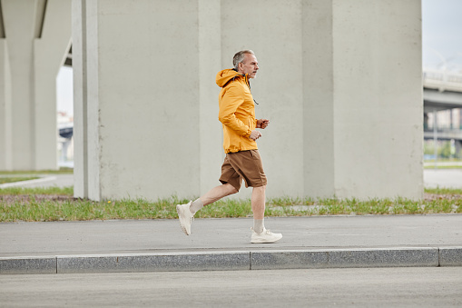 Side view full length portrait of handsome mature man running outdoors in city against concrete background, copy space