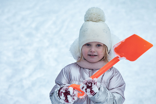 Happy smiling little girl with children shovel in frozen winter snowy playground in warm wear. Cute child having fun walking outdoors. Concept snow wintertime and childhood. Copy text space