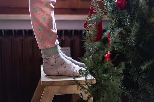 feets in pink socks and pink pajamas of a 7 year old girl who is decorating christmas tree at home during the day timel
