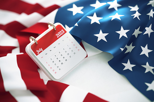 July 2023 calendar over rippled American flag. High angle view and horizontal composition.