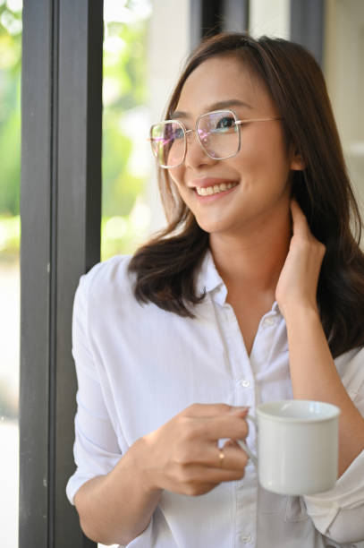 attraente femmina asiatica appoggiata alla finestra, sognando ad occhi aperti mentre si gode il suo caffè. - thai cuisine asian cuisine vertical close up foto e immagini stock