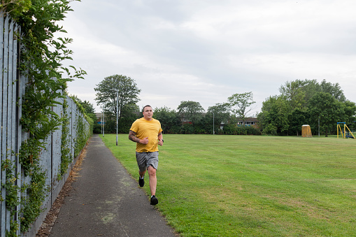 Man doing a workout  near his home in the North East of England. He is jogging through an urban area along a footpath by a field  wearing a smart watch to track his fitness journey.