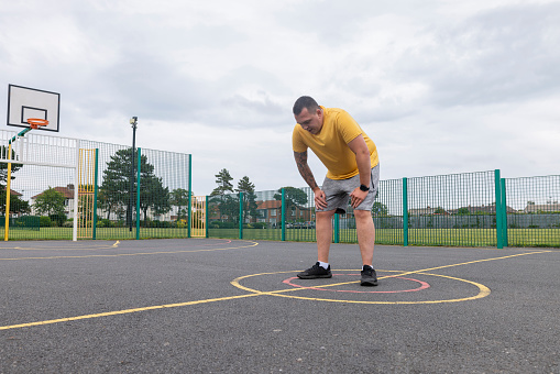 Man doing a workout  near his home in the North East of England. He is catching his breath, bending over with his hands on his knees in a public park on a sports court.