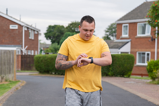 Medium front view shot of a man doing a workout  near his home in the North East of England. He is jogging through an urban area looking down at his smart watch, tracking his fitness journey.