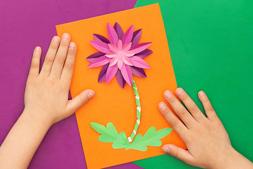 Top view of child hands and papercraft violet crysanthemum flower made by child on orange worksheet on violet and green background. Present for mother s day