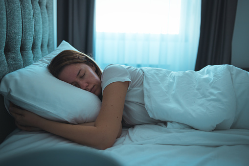 Side view of young beautiful woman dreaming in bed and relaxing at night. High angle view of woman with closed eyes sleeping well at home in the dark. Beautiful girl sleeping peacefully under white blanket in her bedroom at late in night.