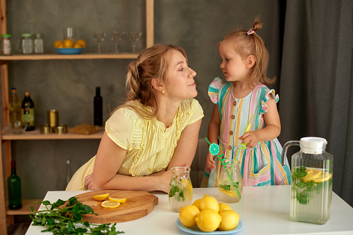 girl with woman drink fresh lemonade in kitchen. mom with love looks at little daughter at home