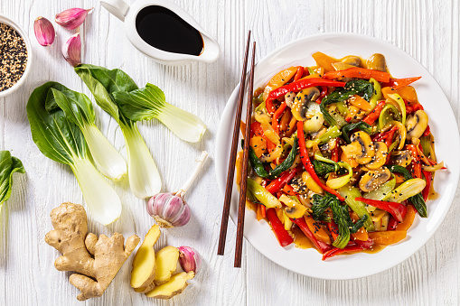 Vegetable Stir Fry of bok choy, red capsicum, mushrooms, onion, carrots and chinese stir fry sauce on white plate on white wooden table with ingredients, horizontal view from above, flat lay