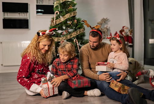 a mother and father with their two small children with Christmas presents in the living room next to the Christmas tree - concept of opening presents -