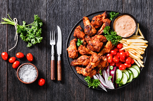 spicy roast chicken wings with french fries, tomatoes, cucumber, red onion and thousand islands dip on black plate on dark wood table, horizontal view from above, flat lay