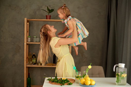 mother and daughter make summer lemon drink at kitchen. woman toss little girl into the air. Happy hugs family time together