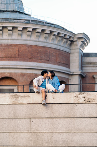 Cool and young asian couple sitting in the city of Madrid and kissing.