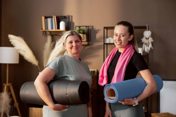 istock Happy elderly mother with young adorable daughter smiling looking at camera holding mat going to work out indoors. In background beautiful modern furniture 1430971236