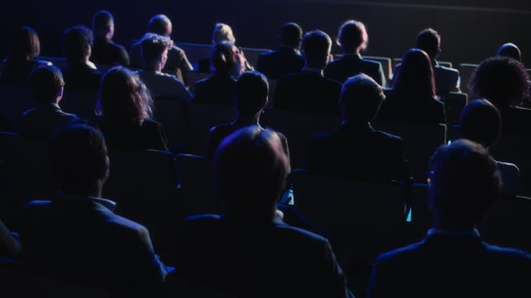 Audience Full of Tech People in Dark Conference Hall Watching an Innovative Inspiring Keynote Presentation. Business Technology Summit Auditorium Room Crowded with Delegates. Static Shot from Behind.