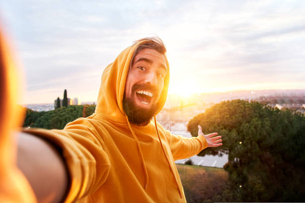 Selfie of young smiling man hooded sweatshirt. Happy Guy with good laugh taking self picture at sunset showing beautiful views. Cheerful traveler enjoying photographing their vacations having fun stock photo
