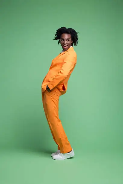 Fashion portrait of elegant man wearing orange suit and white shirt standing with hands in pockets and smiling at camera. Studio shot, green background.