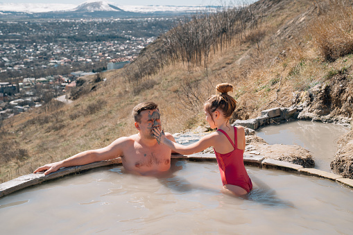 daughter applies healing clay to father's face bathe in hydrogen sulfide thermal spring on mountainside against backdrop of city. therapeutic and relaxing baths in nature at any time of year.