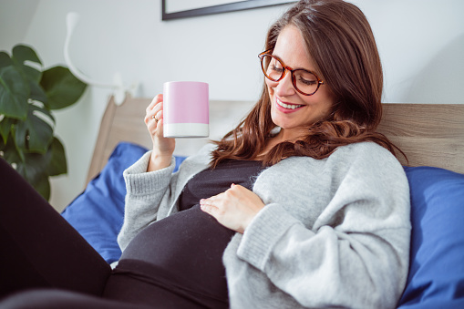 Happy pregnant woman lying on the bed and resting with cup of tea, crouching her belly.