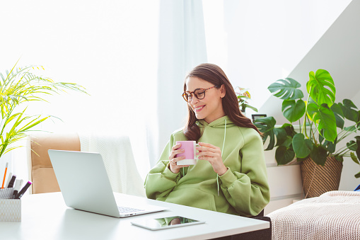 Beautiful woman wearing green hoodie, sitting at the table in her bedroom and having video conference, using laptop, holding cup of coffee. Home office concept.