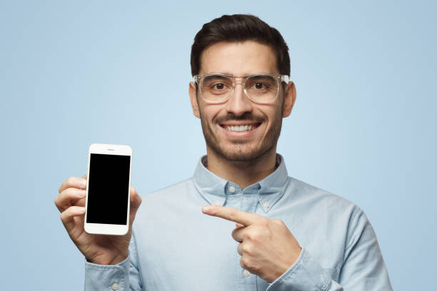 istock Man in shirt on blue background presenting phone and pointing finger at black copy space screen 1430988259