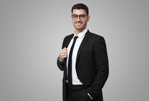Half-length portrait of handsome Caucasian guy dressed in black suit, white shirt and tie isolated on gray background looking attentively through glasses and smiling positively and confidently