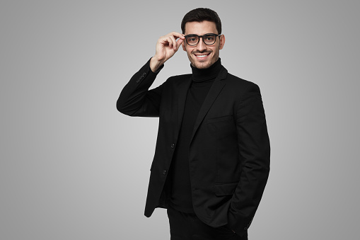 Contemporary intelligent business man wearing black suit holding his spectacles, isolated on grey background
