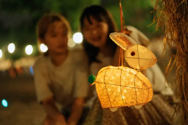 A  traditional Chinese paper rabbit-shaped lantern hung on a tree with its string, as two cheerful young Asian women who are sisters and siblings crouching behind it inside the park, looking together at the lantern, as they have fun and celebrate the Chinese Mid-Autumn Festival together outdoors as a family at night.