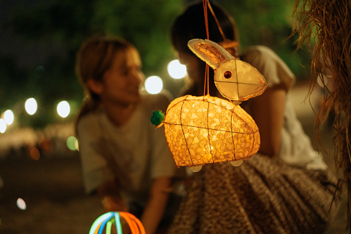A  traditional Chinese paper rabbit-shaped lantern hung on a tree with its string, as two cheerful young Asian women who are sisters and siblings crouching behind it inside the park, looking at each other and talking, as they have fun and celebrate the Chinese Mid-Autumn Festival together outdoors with glow sticks as a family at night.