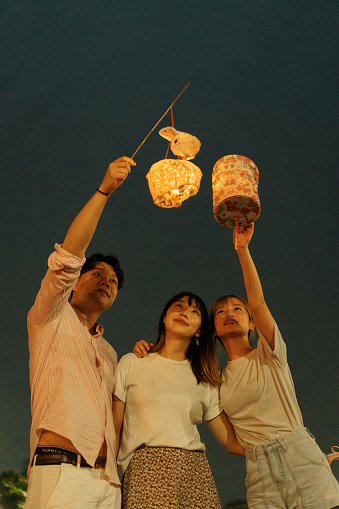 A portrait of a group of two young Asian women and a young Asian man who are friends or family looking at the lit up traditional paper lanterns as they hold them up together and watch the beautiful lights while standing as they celebrate the traditional Chinese Mid-Autumn Festival outdoors at night. Smiling girls and boy enjoying the night view and relaxing around the lights with her friends in a park while they hang out and have fun with the lanterns in the evening in Hong Kong.