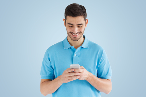 Young smiling man standing isolated on blue background, looking at screen, holding phone with both hands