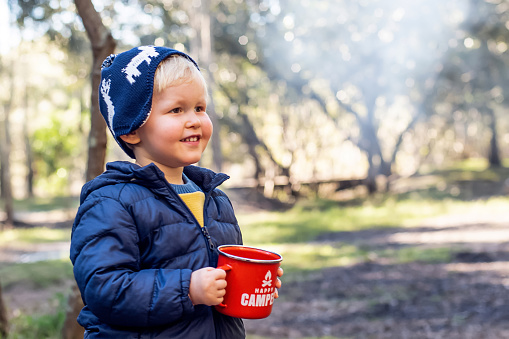 Defocused image of toddler boy on a campsite with the red mug in his hand. Family camping travel with kids concept. Happy camper