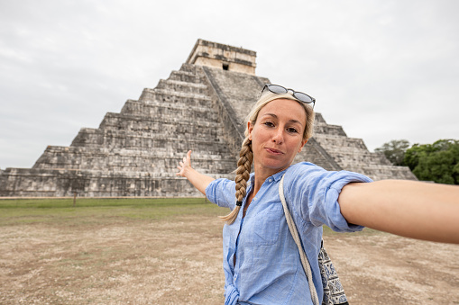 Cheerful female tourist discovering Mayan pyramids in Yucatan, Mexico.
She loves to travel and takes a smily selfie with the ancient temple behind.