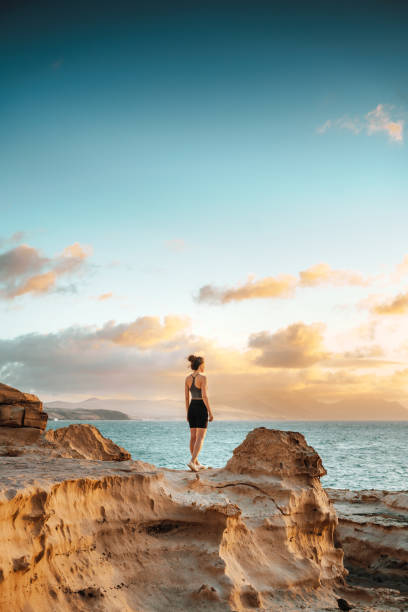 frau steht auf einer klippe mit blick auf den spektakulären sonnenuntergang - fuerteventura stock-fotos und bilder