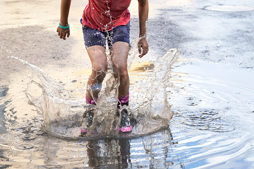 Feet of child in rubber boots jumping over puddle and water splashes in the rain