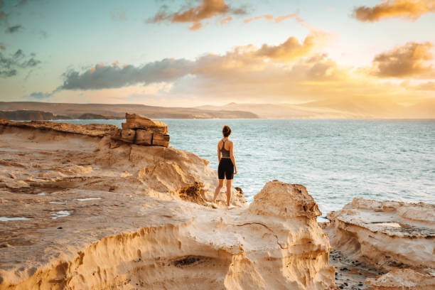 donna in piedi su una scogliera che si affaccia su un tramonto spettacolare - fuerteventura foto e immagini stock