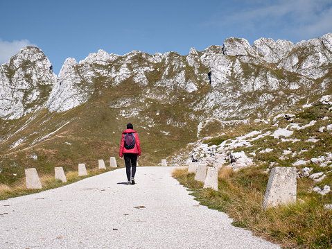 A girl hiking with a backpack among the mountains on an asphalt road in the sun. In the Julian Alps, rocky summits in background