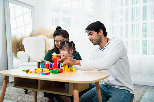 Family sitting together in the living room at house little girl playing with colorful geometric shape toys with her parents watching closely.