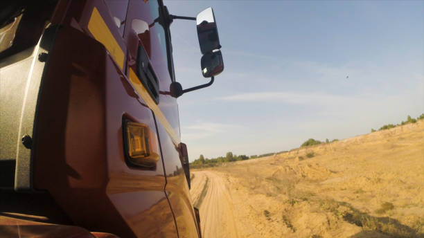 vista laterale della cabina rossa del camion che si muove su una polverosa strada rurale sterrata lungo gli alberi verdi davanti. scena. enorme camion che cavalca in campagna, strada polverosa sullo sfondo del cielo nuvoloso, vista da una ruota. - pick up truck road dust rural scene foto e immagini stock