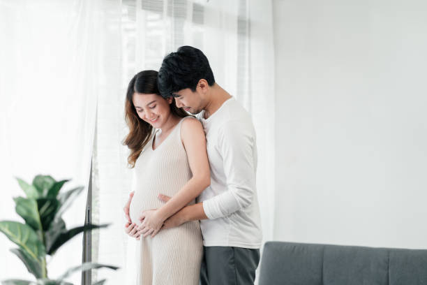 A handsome husband is  embracing his beautiful pregnant wife next to the window in the living room. stock photo