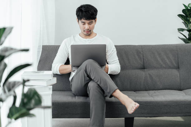 el apuesto hombre asiático independiente está trabajando en línea en una computadora portátil en la sala de estar de su casa. - con las piernas cruzadas fotografías e imágenes de stock