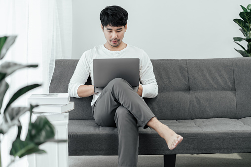 Handsome Asian man freelance is working online on computer laptop in the living room at home. The concept of work from home. Young male college searching for studying or shopping online at home.