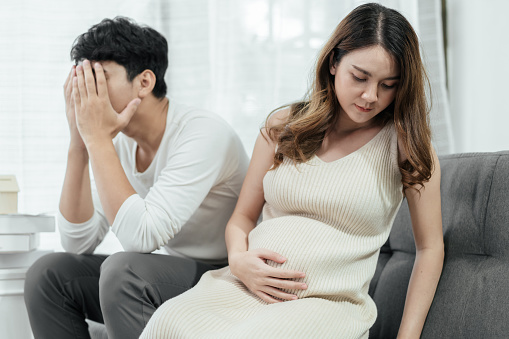 Pregnant asian couple seem stressed, the husband's hands hold his head, stressfully. His wife turns her back on him with concern on her face. The concept of family issues.
