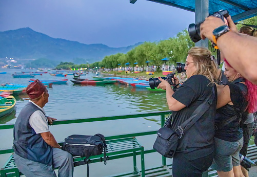 Pokhara, Nepal - April 21, 2022:  A group of tourist at Phewa lake. They all have a camera, taking photographs of a serene Nepali man wearing the traditional hat, relaxing on a bench.