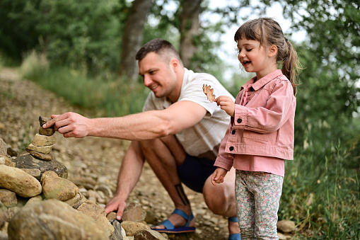 oddler playing with a dry leaf while her dad try to build tower of stones in a woodland countryside nature forest outdoors. happy family outdoors lifestyle. father with smiling daughter playing in a game with stones.Family builds stone stacks.
