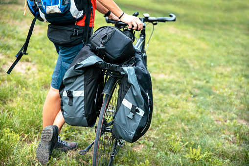Back of Unrecognizable Woman backpacker with bags on bicycle walking and pushing her bike on the green grass outdoor. Traveling, cycling, tourist, economic travel concept. Copy space